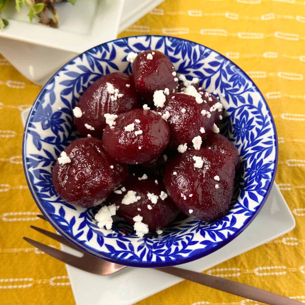 Whole pickled beets garnished with goat cheese crumbles in a decorative blue bowl.