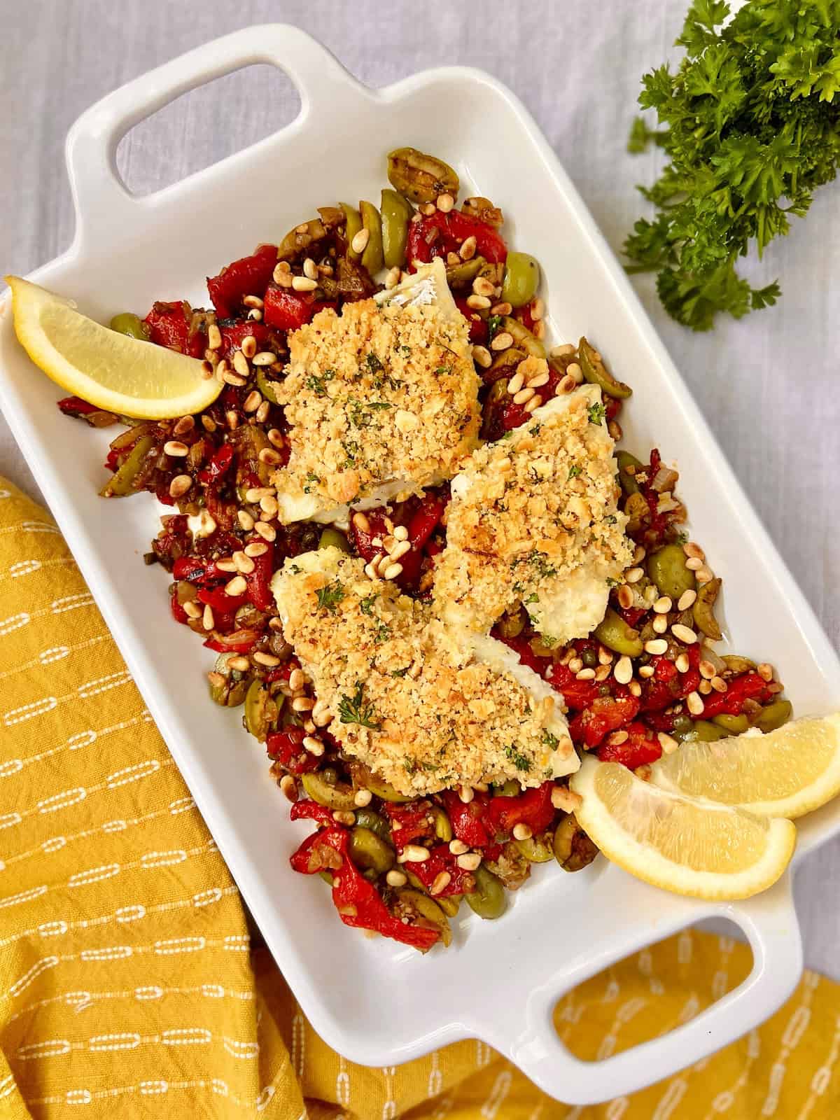 Sicilian Baked Cod in white serving platter garnished with red peppers, olives and lemons.