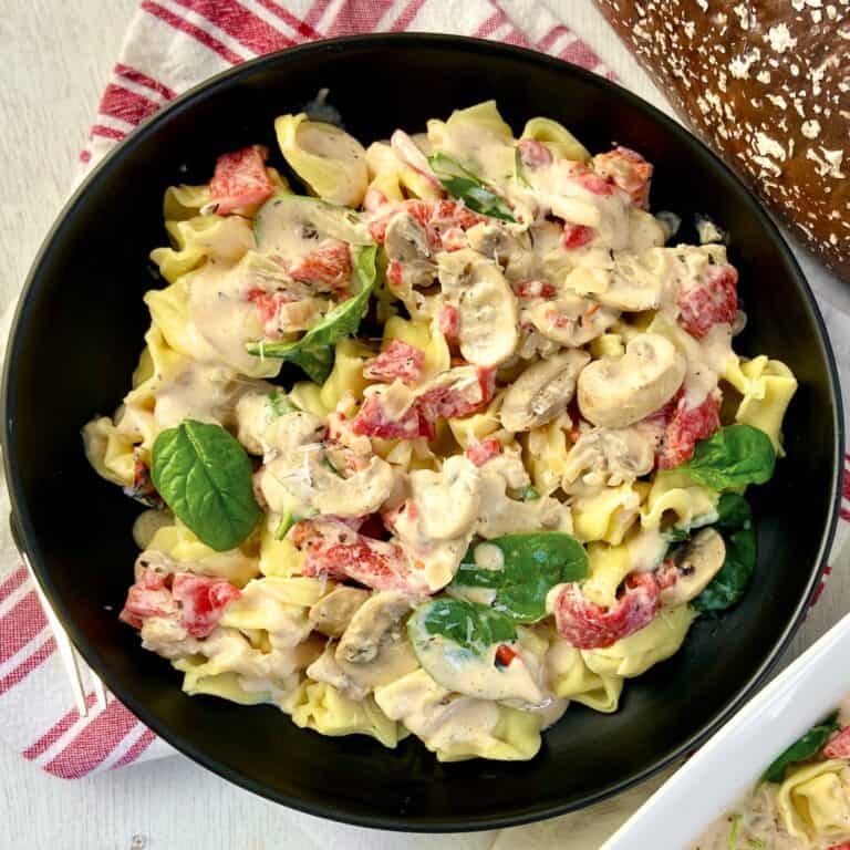 Tuscan cream sauce over tortellini in black bowl with roasted red peppers, mushrooms and spinach.