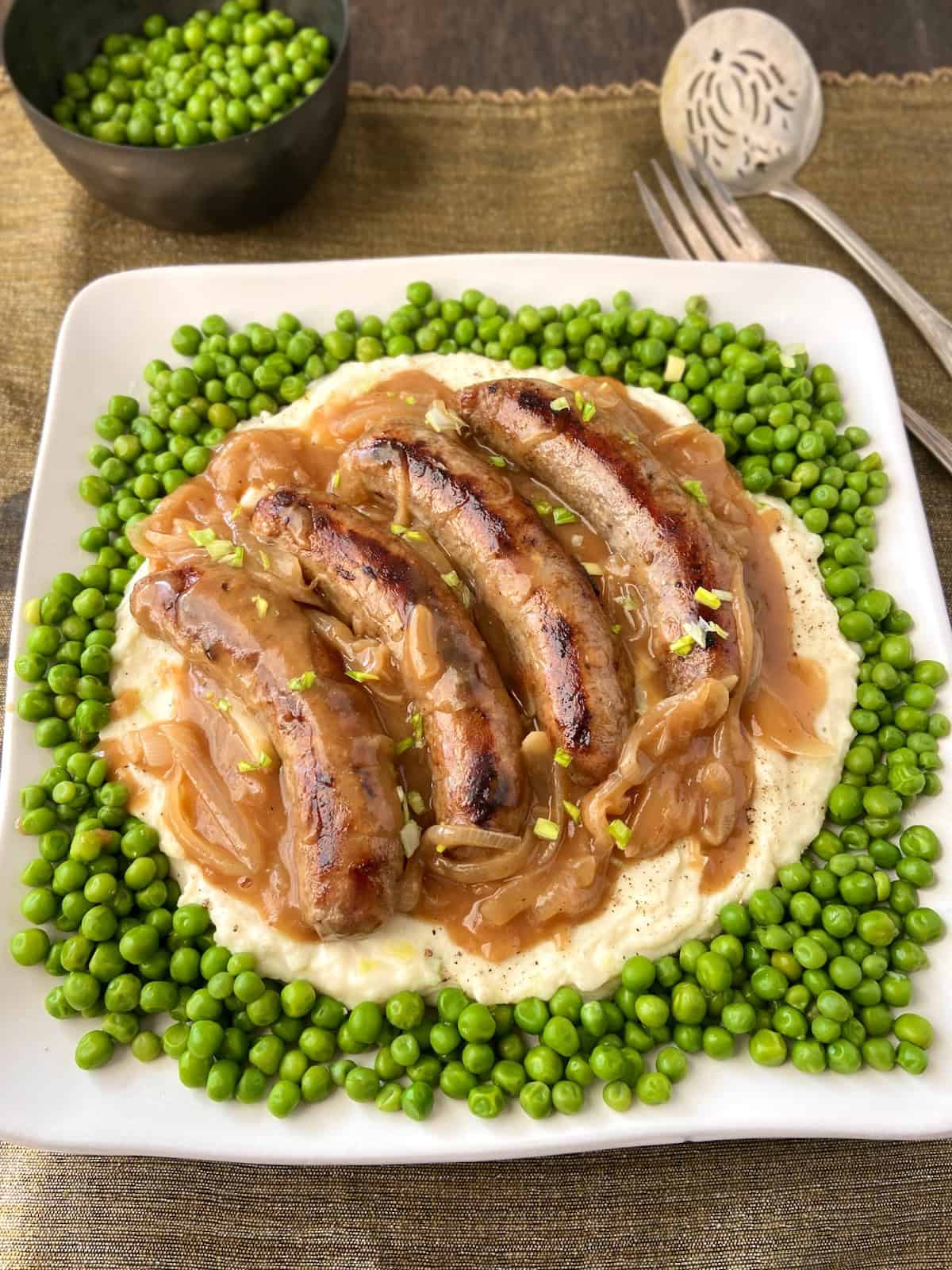 Bangers, mash, onion gravy and green peas on white plate.